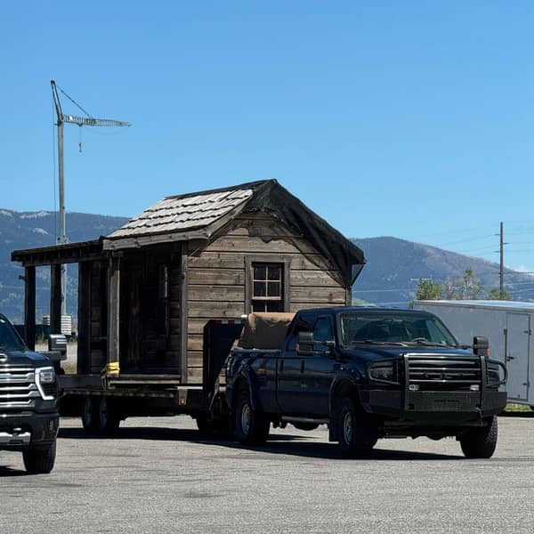 Dark pickup towing a small wooden cabin on a flatbed trailer